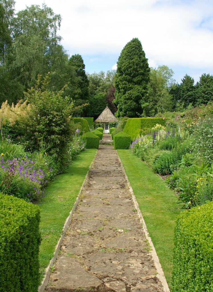 A view down a garden path flanked by clipped hedges and full borders to the gazebo beyond.