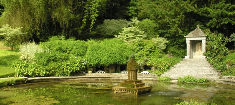 A view across the pond and fountain leading to a stepped gazebo surrounded by plants and shrubs.