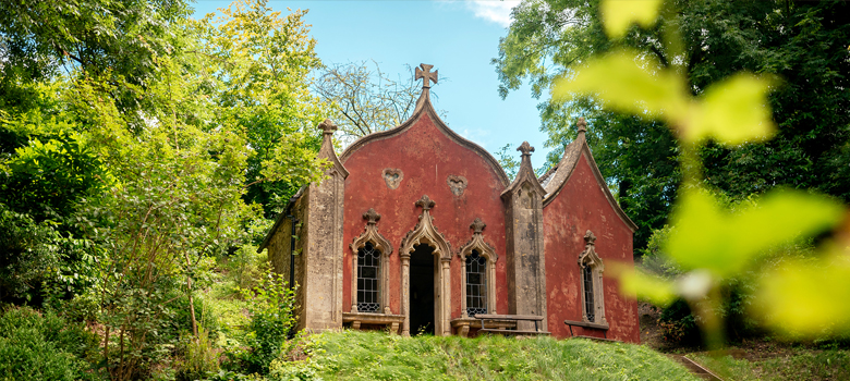 A view through trees of one of the rococo style buildings above a slope at Painswick Rococo Gardens.